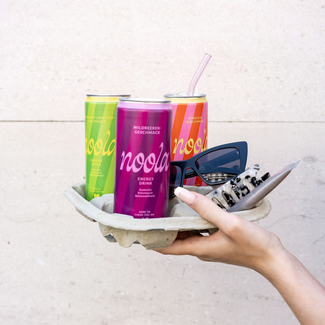 3 noola Energy Drink Dosen in Wildbeere, Pfirsich und Zitrone-Limette werden in einem Papptray von einer Hand gehalten