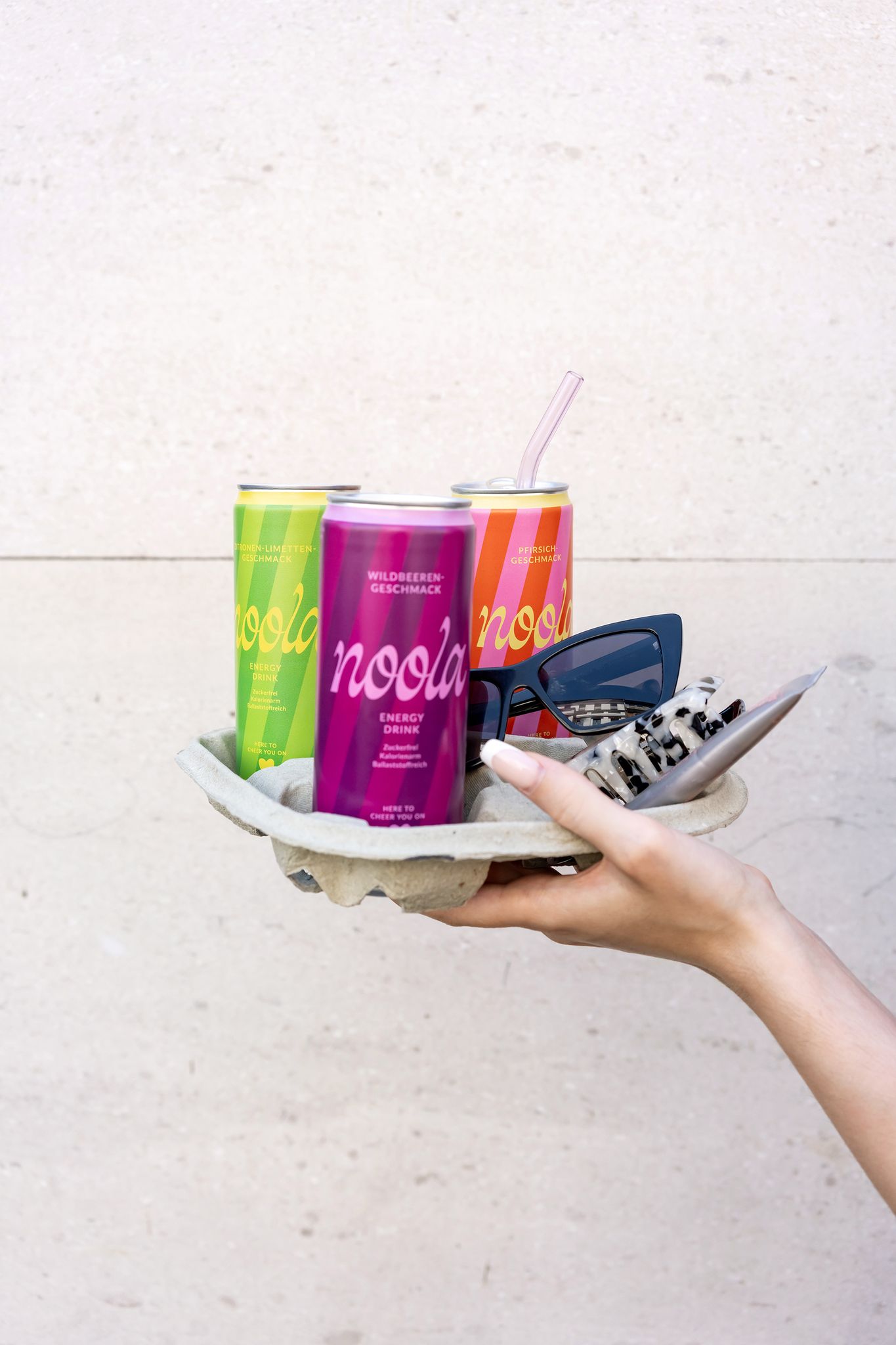 3 noola Energy Drink Dosen in Wildbeere, Pfirsich und Zitrone-Limette werden in einem Papptray von einer Hand gehalten